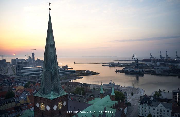 Aarhus Domkirke med spiret i forgrunden. Solen er ved at stå op, og i baggrunden står solen op ved Aarhus Ø. Rosa og irgrønne farver.