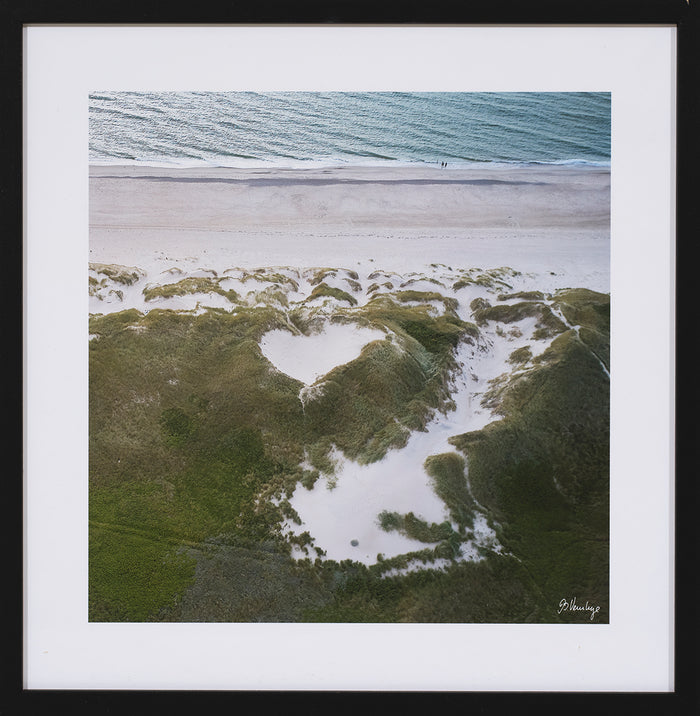 Klitterne i Hvide sande. Hav, skumsprøjt, og en gryde i klitterne der ligner et hjerte. Grøn sand og lyseblå farver.