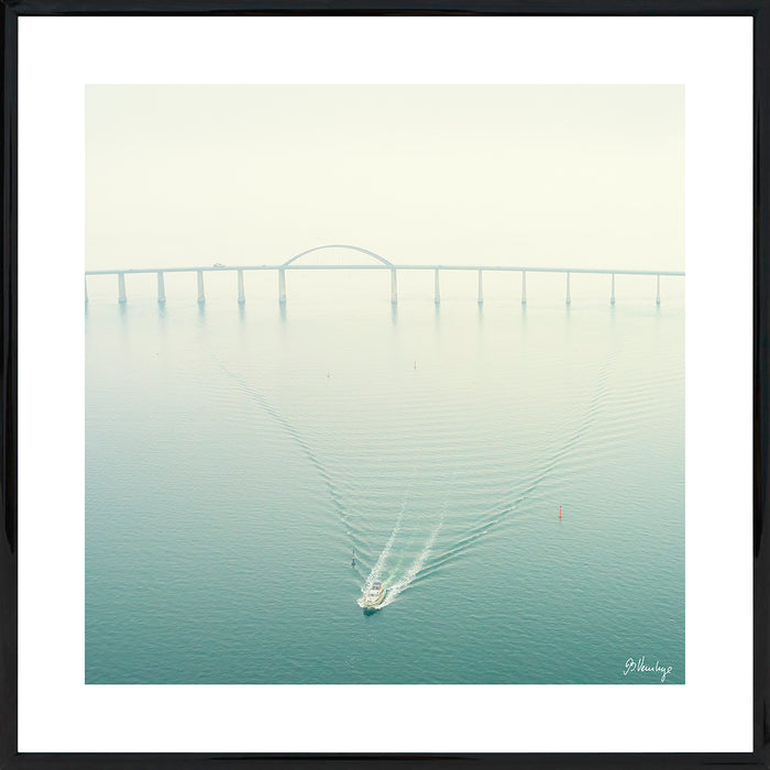 Langelandsbroen på en diset dag. En mindre båd trækker mønstre i vandet. Sart tyrkisgrøn med en rød bøje.